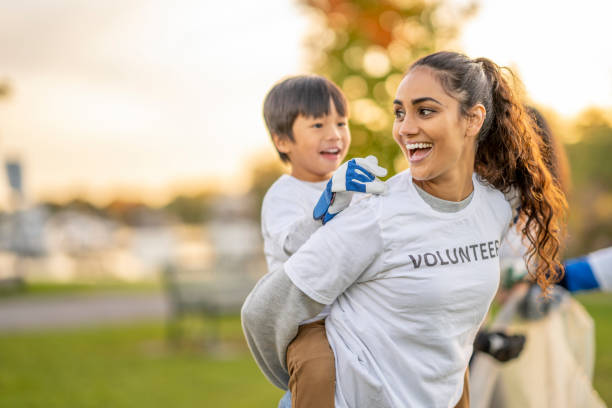 Two young community volunteers are seen enjoying their time together outside.  The woman is giving the young boy a piggyback ride as they laugh and enjoy the day.